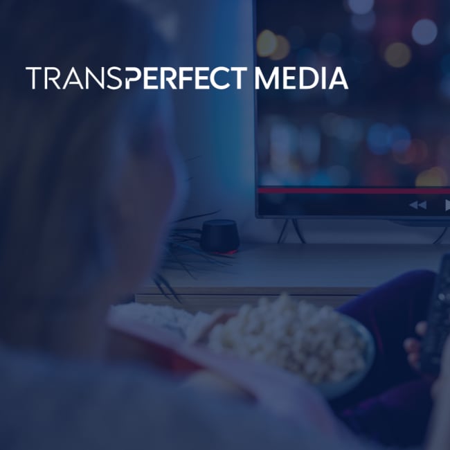 A woman sits on her couch with popcorn and a tv remote pointed at her tv while she pauses/plays a film playing. "TransPerfect Media" logo in the corner of image.